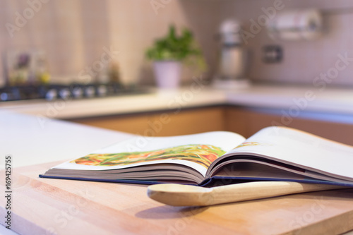 Cook book put on a kitchen table with a wooden spoon in the foreground. Kitchen is visible in the background. photo