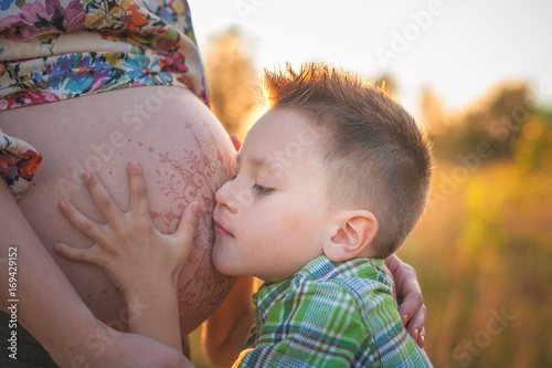 Cute boy kissing and listening his mothers pregnant mehandi pattern ornament belly on sunset nature landscape