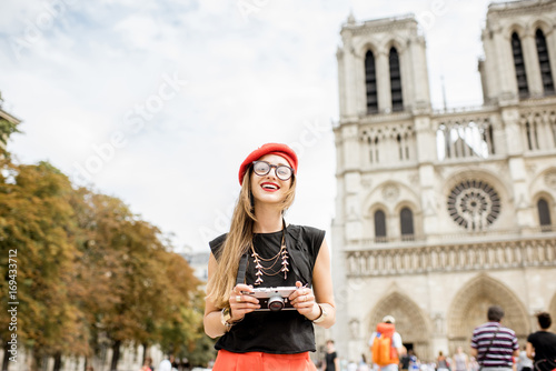 Young woman tourist in red cap standing with photo camera in front of the famous Notre Dame cathedral in Paris