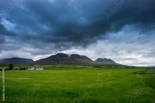 Iceland - Green meadows and fields before snowy volcanoes in dark cloudscape of thunderstorm