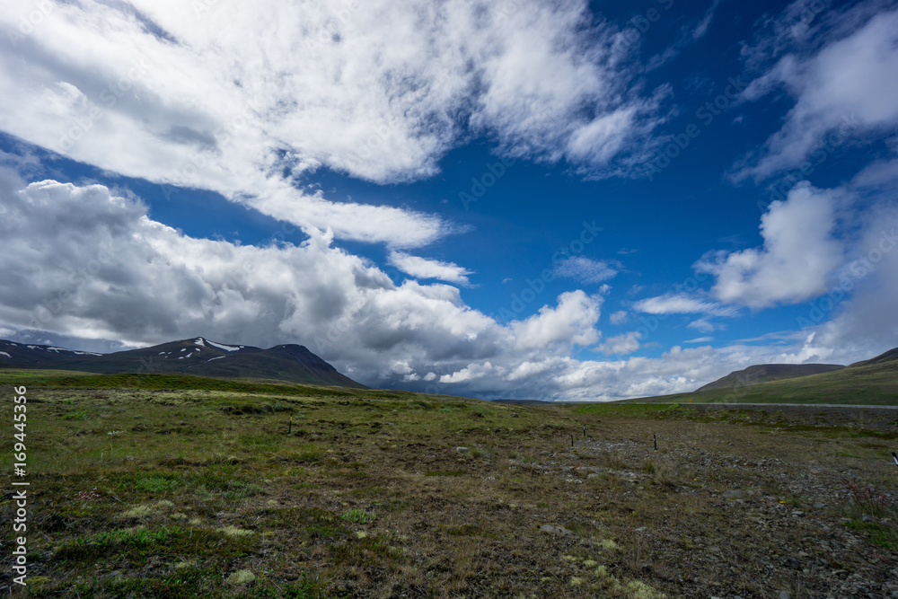 Iceland - Blue sky with sun and dark clouds with thunderstorm coming over green landscape