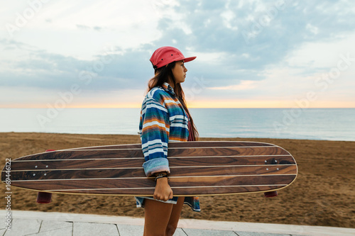 Side view of a trendy girl holding her longboard by the beach at sunrise.