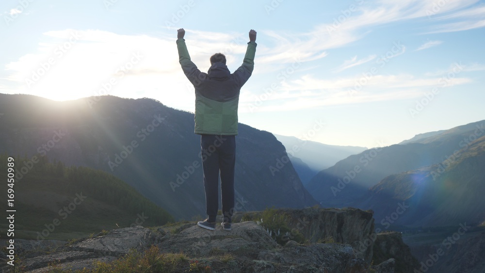 Success young man raising his hands high on top of the big mountain at beautiful golden sunset