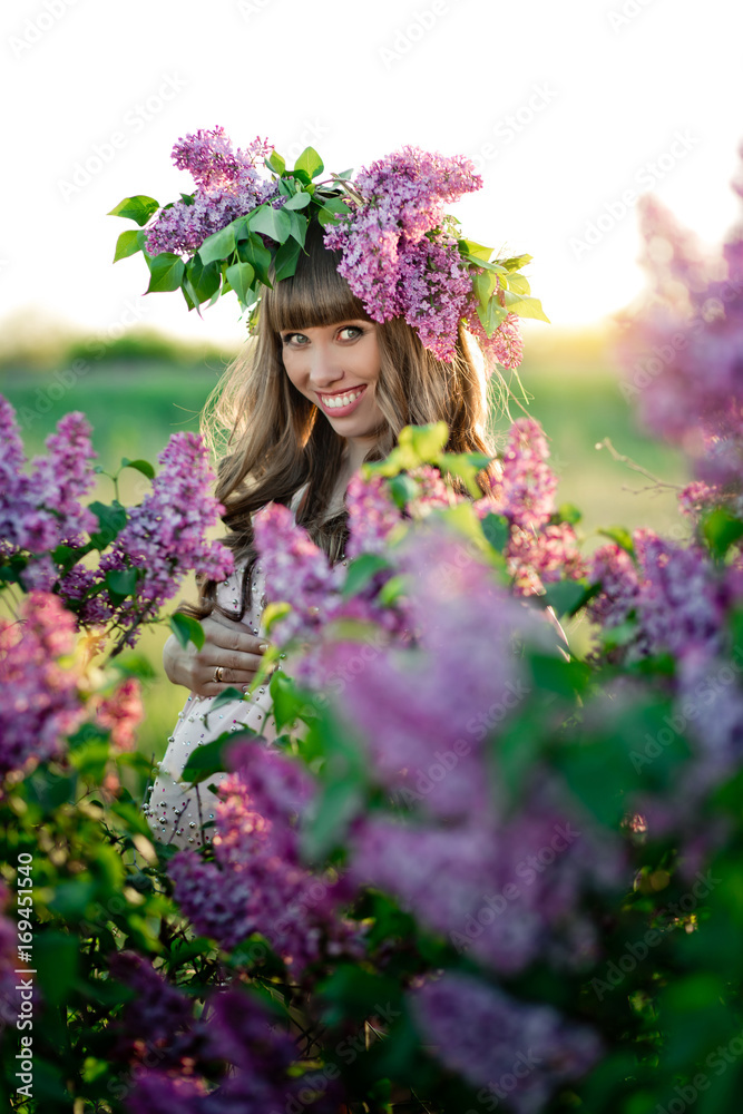The young pregnant woman with a wreath on her head is looking through a lilac bush The young pregnant woman with a wreath on her head is looking through a lilac bush