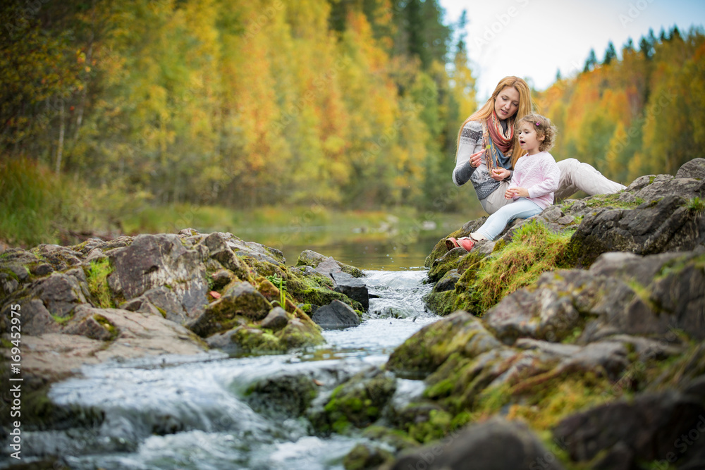 Sweet, cute little girl and mommy sitting on a rock in forest at stream. Enjoying fresh autumn air. Yellow trees. Karelia. Waterfall Kivach.