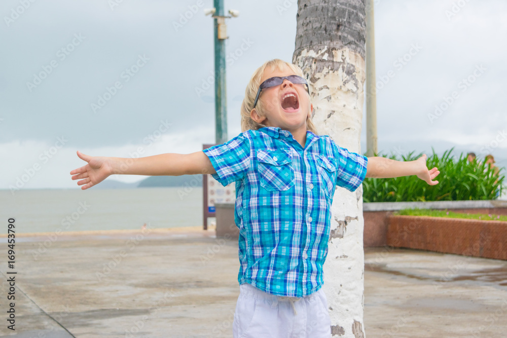 A cool blond boy in sunglasses screaming happyly in a park near beach