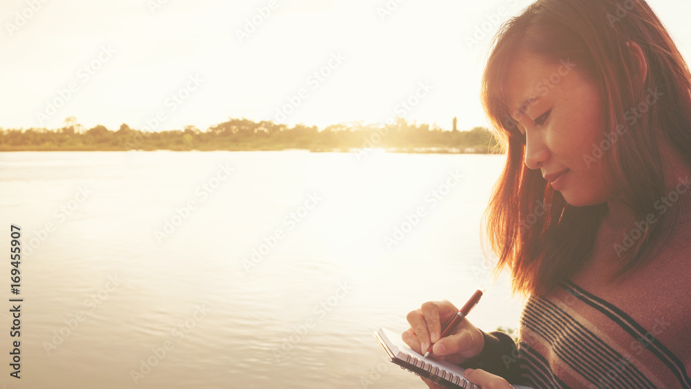 Young woman with pen writing on notebook at riverside in the evening.