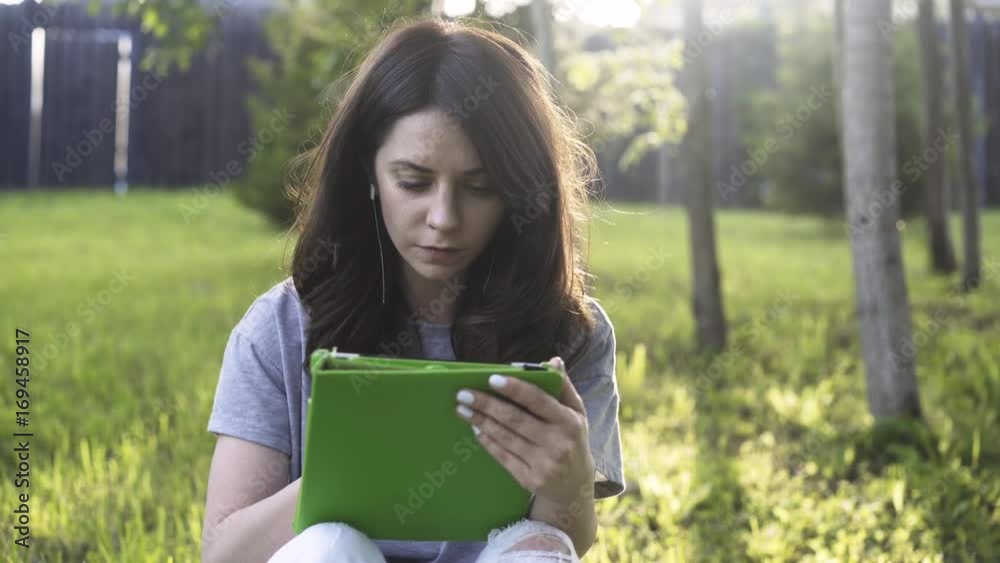 Close up of a young woman wearing a gray T shirt with her tablet computer on a lawn in a park on a sunny day. Left to right pan lear time medium shot Close up of a young woman wearing a gray T shirt with her tablet computer on a lawn in a park on a sunny day. Left to right pan lear time medium shot