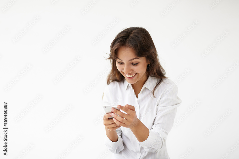 Positive human reaction, attitude and modern technology. Good-looking cheerful young Hispanic businesswoman or office worker in white formal wear reading sms on cell phone and smiling broadly Positive human reaction, attitude and modern technology. Good-looking cheerful young Hispanic businesswoman or office worker in white formal wear reading sms on cell phone and smiling broadly