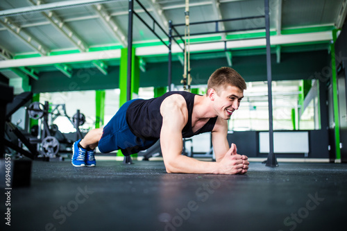Fototapeta Naklejka Na Ścianę i Meble -  Confident muscled young man wearing sport wear and doing plank position while exercising on the gym interior