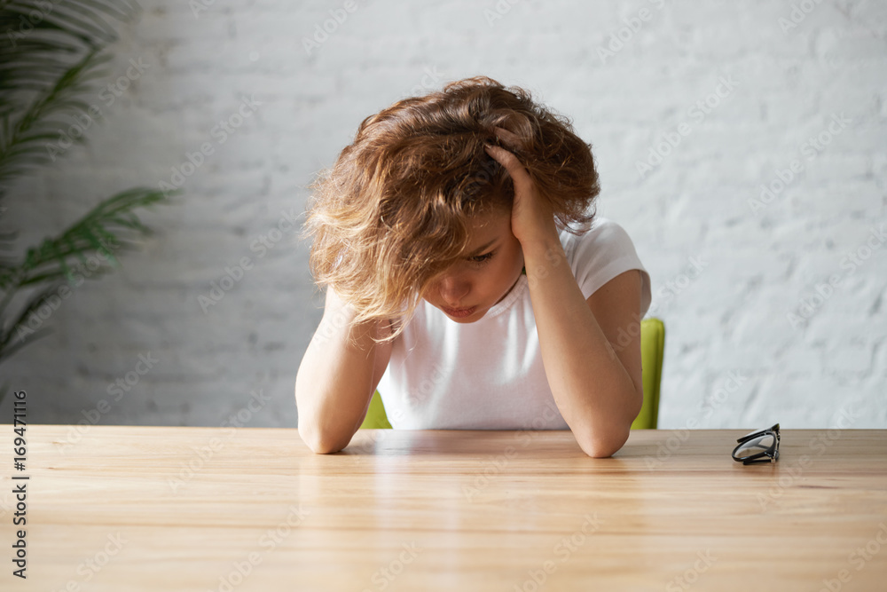 Under pressure of problems. Picture of sad and despressed young Caucasian female sitting at table, holding hands on her head, feeling frustrated while facing financial troubles or stress at work Under pressure of problems. Picture of sad and despressed young Caucasian female sitting at table, holding hands on her head, feeling frustrated while facing financial troubles or stress at work
