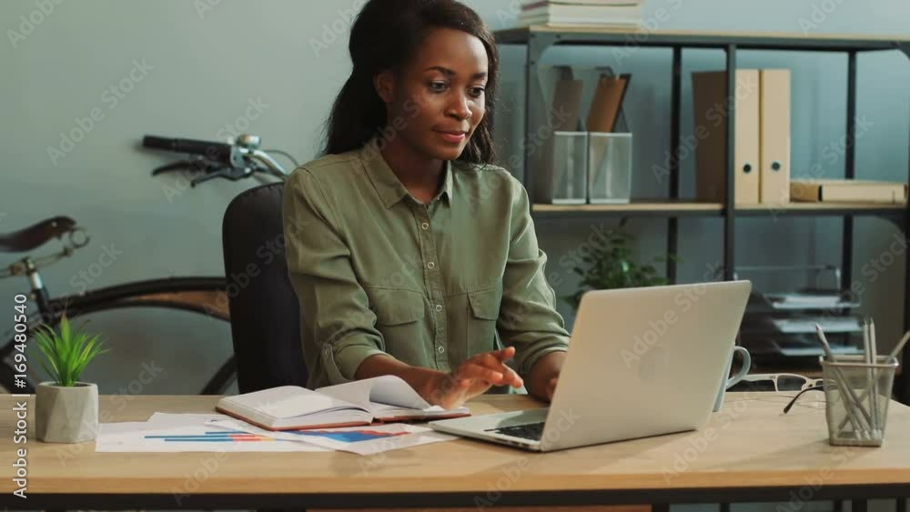 Young attractive african business woman finishing finance report on the laptop in the modern office and feeling happy. Young attractive african business woman finishing finance report on the laptop in the modern office and feeling happy.
