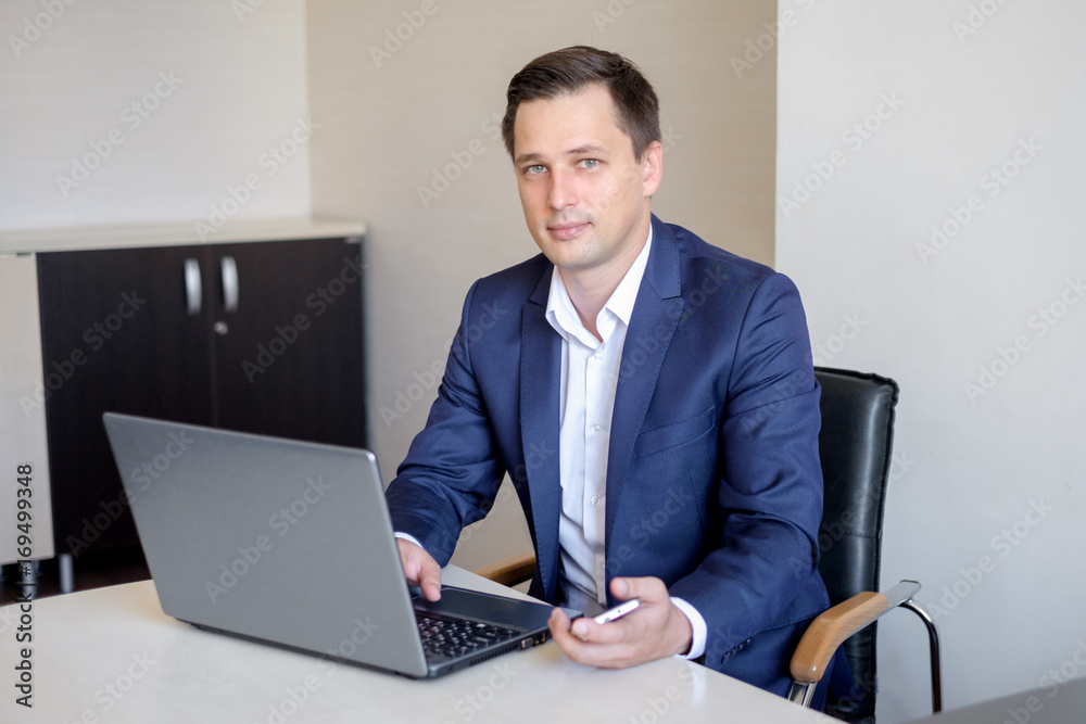 Smiling young businessman sitting behind his desk with laptop and talking on mobile phone in office Smiling young businessman sitting behind his desk with laptop and talking on mobile phone in office