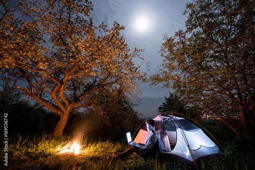 Female tourist using her laptop in the camping at night. Woman sitting near campfire and tent under trees and night sky with the moon