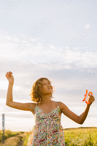 Girl in dress holding bright kite while running and looking away in green summer field. © kkolosov