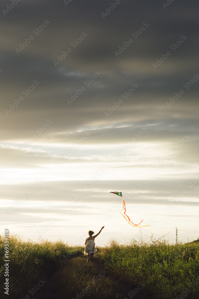 Fototapeta premium Back view of girl holding kite and running in field under sky with overcast.