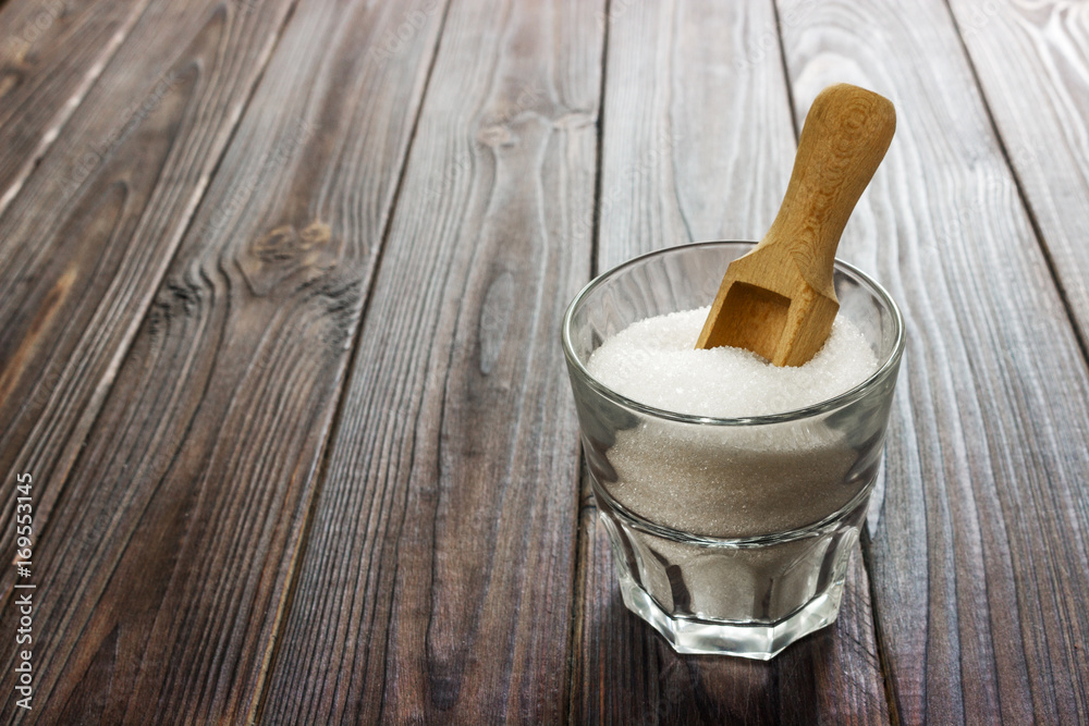 White granulated sugar in white cup with wooden spoon on wooden background.