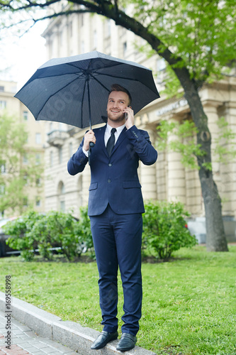 Well-dressed employee speaking on smartphone under umbrella
