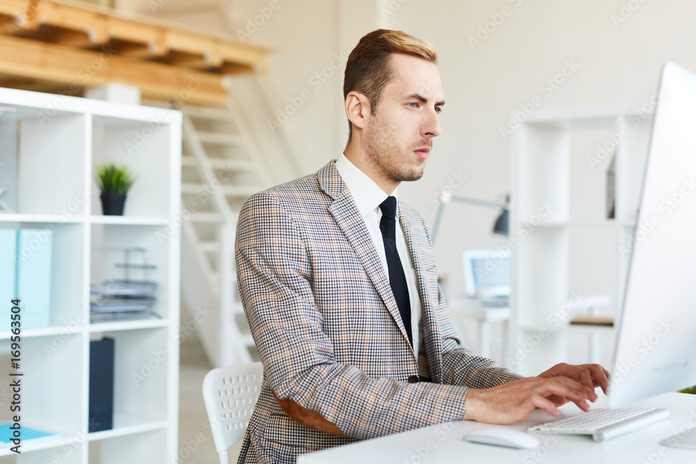 Young businessman typing while looking at computer monitor by workplace