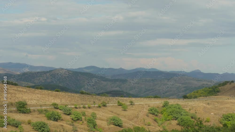 Suburban road through hilly terrain / Road through hilly terrain on background of blue mountains and gray clouds Suburban road through hilly terrain / Road through hilly terrain on background of blue mountains and gray clouds