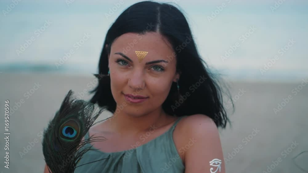 Outdoor fashion portrait of beautiful brunette lady at beach with flash tattoos on hands, back and forehead. Gypsy boho style. Woman in long green dress with peacock fan. Slow motion.