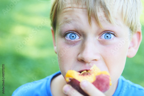 Portrait of child boy eating peach. Happy child in sun summer day. Kid with fruit in nature background.