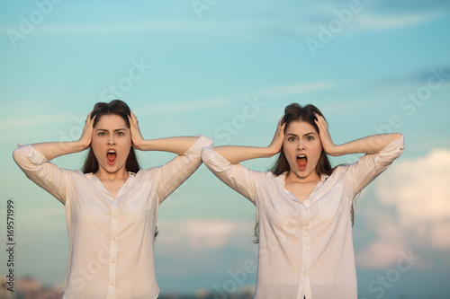 Two beautiful young girl sisters twins on blue sky background with frightened facial expressions on face