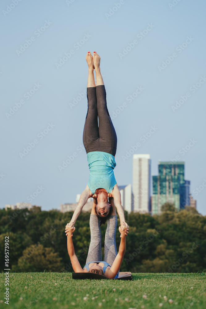 Two young Caucasian women yogi doing star shoulder stand acro yoga pose.  Women doing stretching workout in park outdoors at sunset. Healthy  lifestyle modern activity Stock Photo