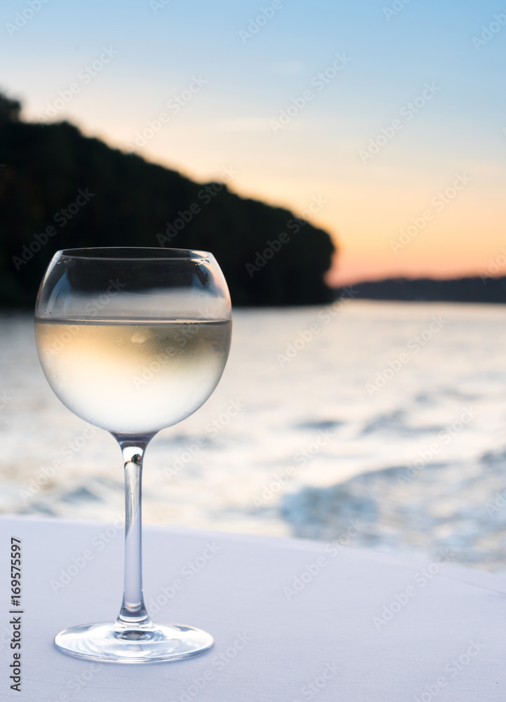glass of cool white wine on white tablecloth with coastline in blurred background at sunset