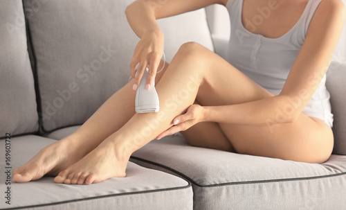Foto Beautiful young woman epilating legs while sitting on sofa at home