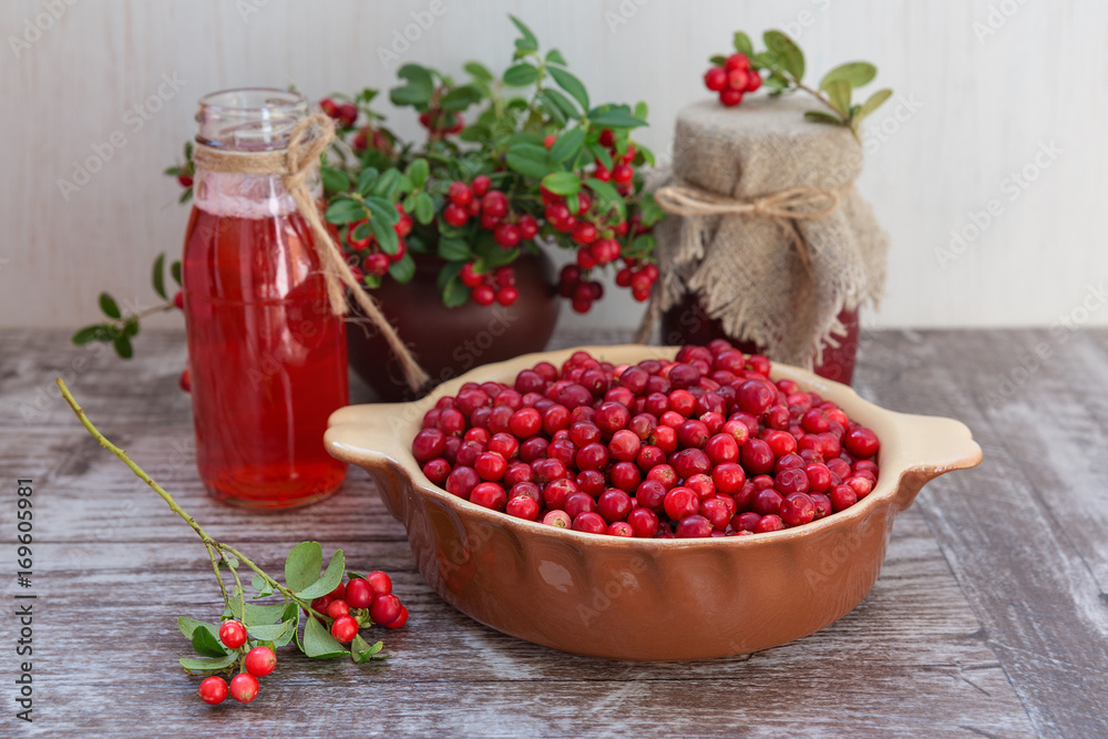 Very ripe cowberries in a bowl on the table and cranberry drinks and jam. The bouquet of sprigs of cranberries in a pot. On a wooden background, space for text. Very ripe cowberries in a bowl on the table and cranberry drinks and jam. The bouquet of sprigs of cranberries in a pot. On a wooden background, space for text.