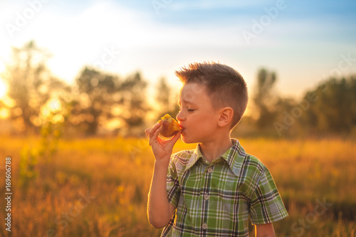 Portrait of young boy eating peach. Happy child in sun summer day. Kid with fruit in nature background at sunset.