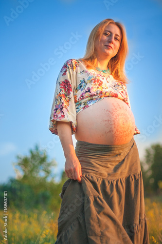 9 months pregnant woman with mehandi pattern ornament belly standing in yellow grass and smiling on sunset lights. Waiting for baby. Pregnancy concept