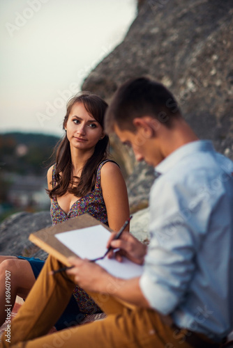 Young lovely couple on a romantic open-air date. A young man paints a portrait of his beloved girl sitting on a rock.
