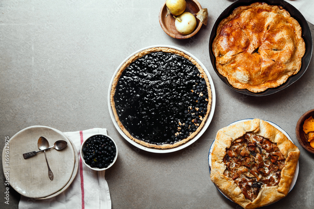 Variety of autumn pies: blueberry tart, chantarelle galette and classical apple pie with sweet pastry crust. Harvest time baking concept. Concrete rustic background. Space for text.