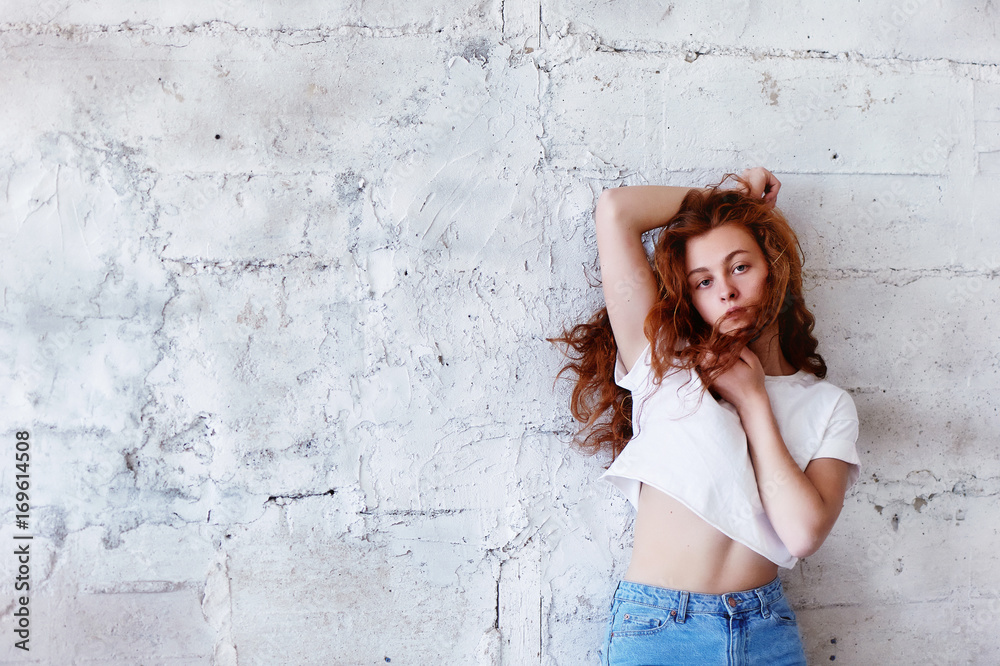Model tests. Beautiful redhead girl with curly hair. Natural color. Stands with long hair blowing against the light brick wall. Studio-loft Model tests. Beautiful redhead girl with curly hair. Natural color. Stands with long hair blowing against the light brick wall. Studio-loft