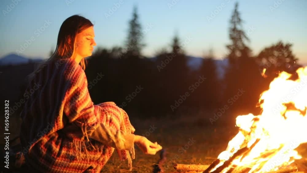Side view mountains woman girl sitting wrapped blanket camp fire putting firewood burning wood branch evening dusk camping sunset closeup orange red flame warm hot color wild nature burn cozy silence
