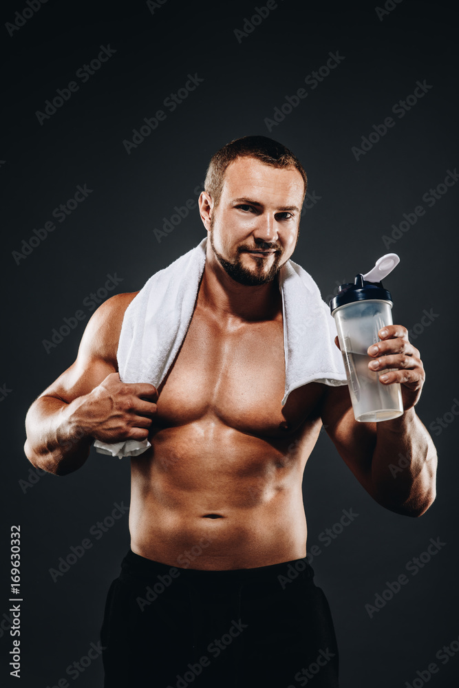 A handsome young sportsman standing half naked with a towel in his neck , Bodybuilder after a workout with a shaker on  dark background