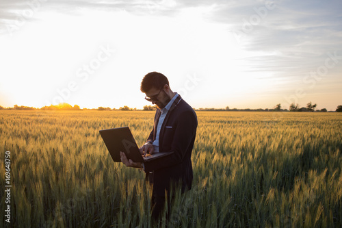Young man in black suit using laptop in wheat field, during beautiful sunset 