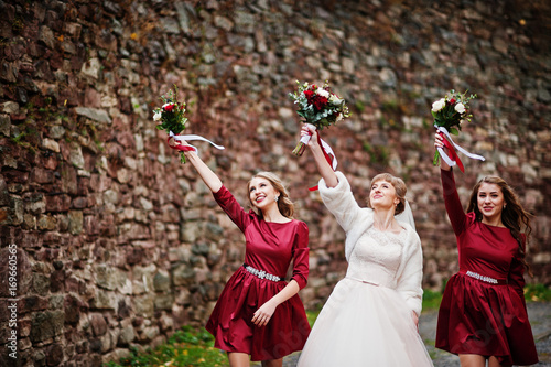 Magnificent bride walking and posing with her bridesmaids with bouquets in the park next to the old brick wall.