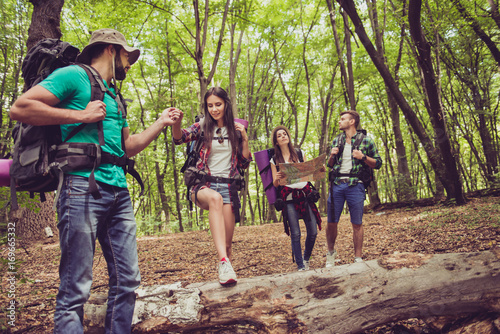 Cute young brunette is stepping over huge log, the bearded guy her lover is holding her hand, helping during hiking. Other couple looks around, holding map. Four tourists best friends © deagreez