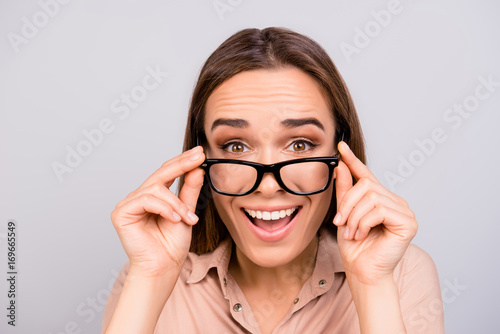Close up of an amazed brunette young woman in beige formal shirt and glasses, holding them, she is shocked, extremely happy, with wide open eyes, on pure light grey background
