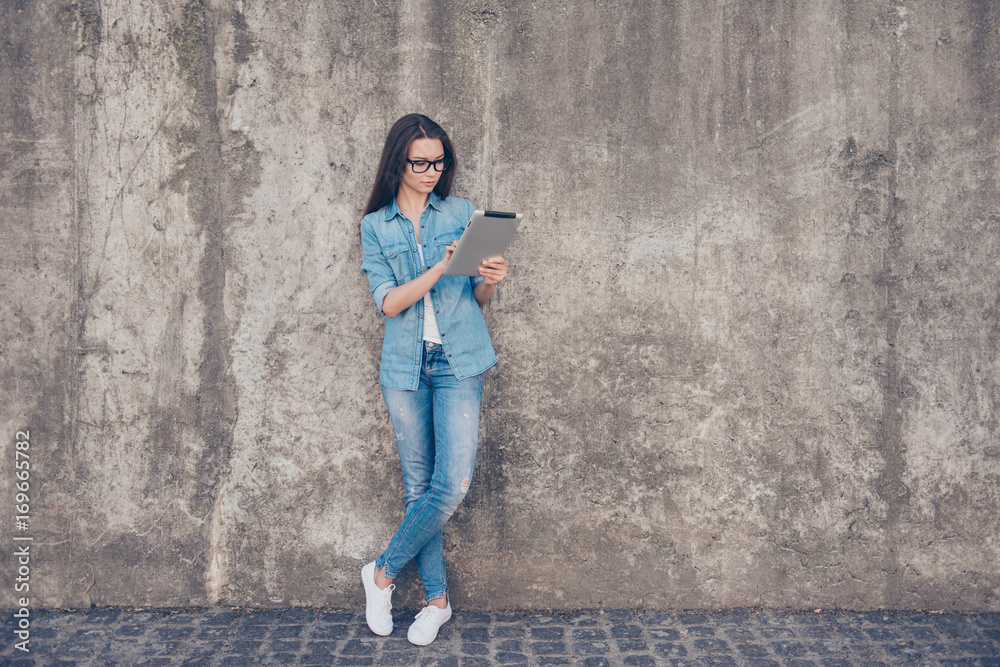 Success and work, study and business concept. Young lady is working on her tablet, wearing casual jeans clothes and glasses near concrete wall outdoors with crossed legs