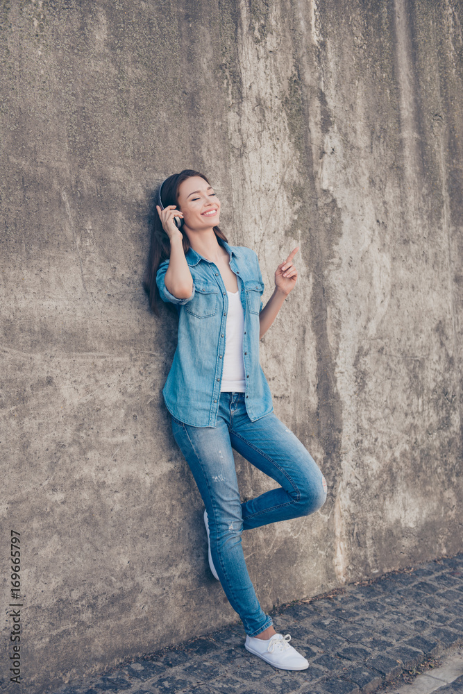 Enjoy the music! Young cute hipster is leaning the concrete wall in the city, listening to the music, gesturing, having fun, in modern denim outfit