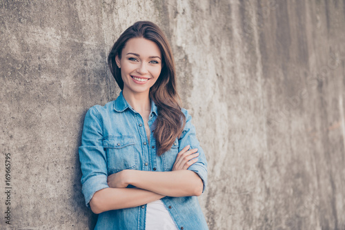 Adorable young brunette lady is leaning the concrete wall outdoors, smiling, with crossed arms, in casual denim jeans outfit