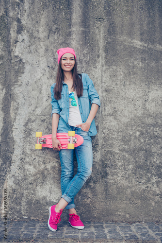 Youth culture. Young attractive hipster girl stands with skate board on concrete wall`s background, in denim outfit, pink hat and shoes, holds a long board