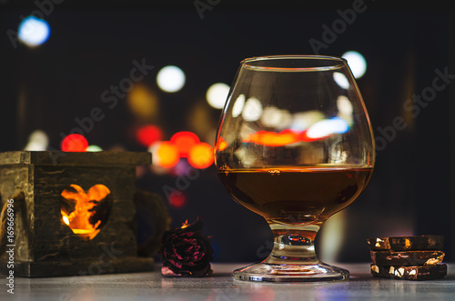 Close-up view of glass with brandy or cognac, chocolate and candle on color background with night city lights