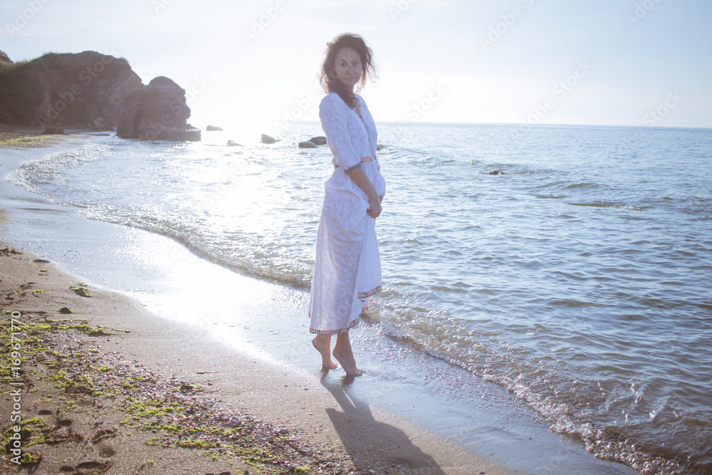 Young brunette woman posing on the morning summer beach in beautiful white dress Young brunette woman posing on the morning summer beach in beautiful white dress