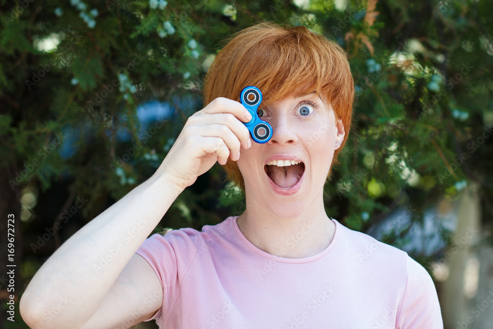 smilling girl in pink t-shirt is playing blue metal spinner in hands on the street, woman playing with a popular fidget spinner toy, anxiety relief toy, anti stress and relaxation fidgets