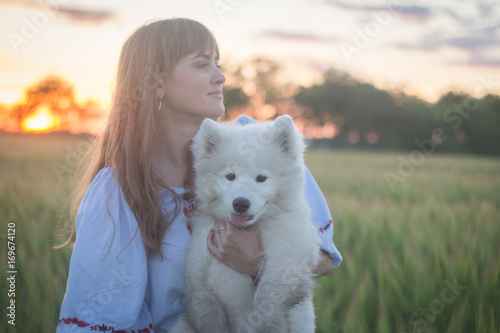 portrait of woman and white puppy of husky dog in the fields, green grass and sunset background © serejkakovalev